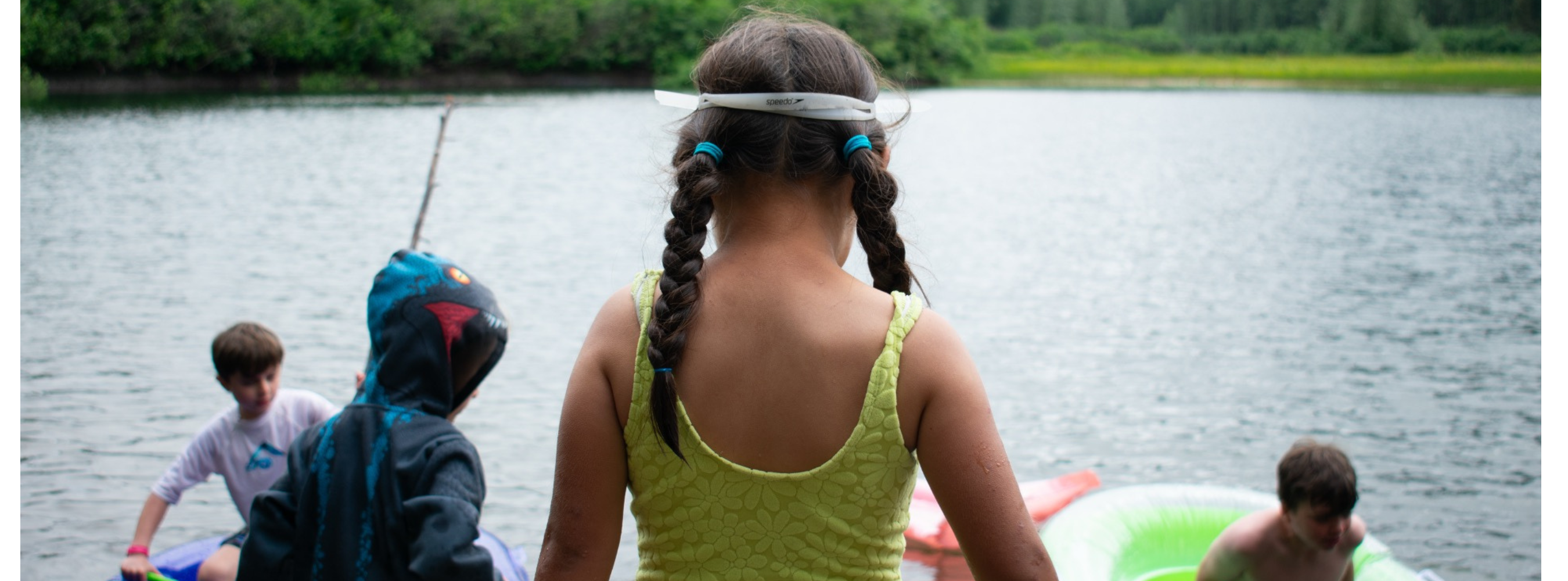 A girl confidently walks towards a waterfront as other youth enjoy the water with inflatable water gear