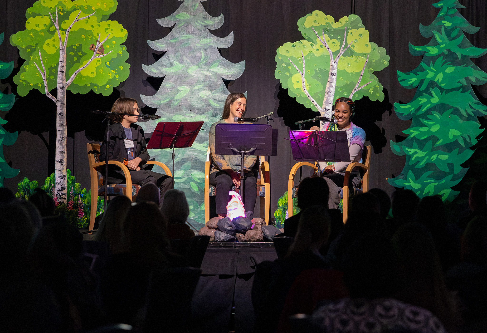 Three people sitting around an artificial camp fire, on a stage. Painted wooden trees are behind them. 