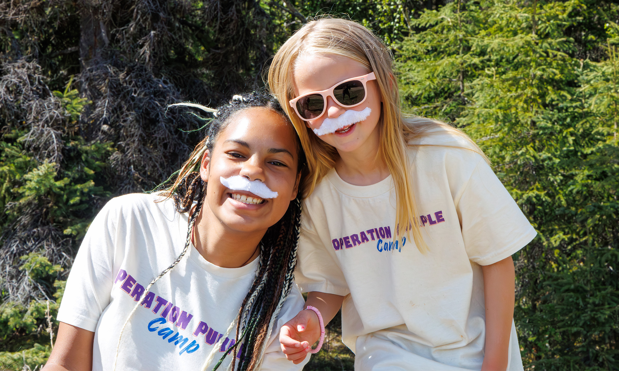 A counselor with a camper on a sunny day. Both are wearing white, fuzzy moustaches.
