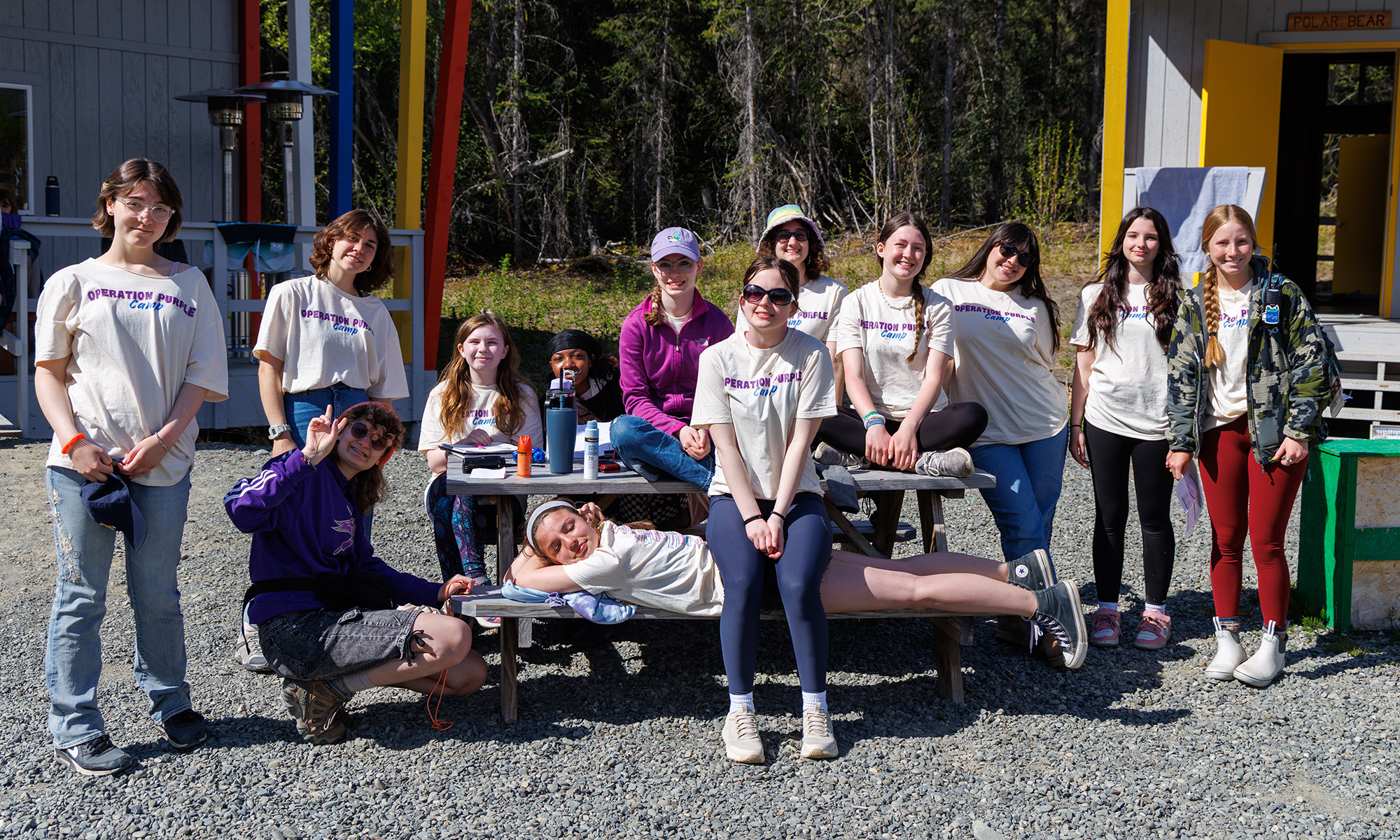 A group of 13 teen campers posting around a picnic table.