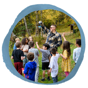 Image in circle frame overlayed on a piece of blue paper. The image shows an adult holding up natural items in a small clear case while a group of kids raise their hands. Fall foliage is behind them.